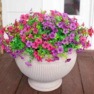 May include: A large, off-white, fluted pedestal planter filled with a dense arrangement of artificial flowers in vibrant shades of magenta, purple, and violet, accented by small, bright green leaves that spill over the rounded rim of the pot.