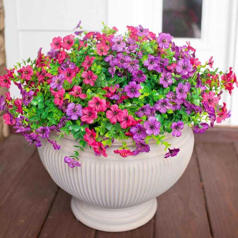 May include: A large, off-white, fluted pedestal planter filled with a dense arrangement of artificial flowers in vibrant shades of magenta, purple, and violet, accented by small, bright green leaves that spill over the rounded rim of the pot.