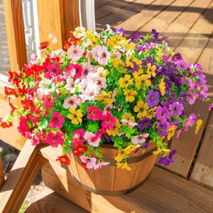 May include: A wooden barrel planter filled with a dense arrangement of artificial flowers in vibrant shades of red, pink, yellow, and purple. The flowers feature small, five-petalled blossoms with green foliage, creating a full, rounded display.