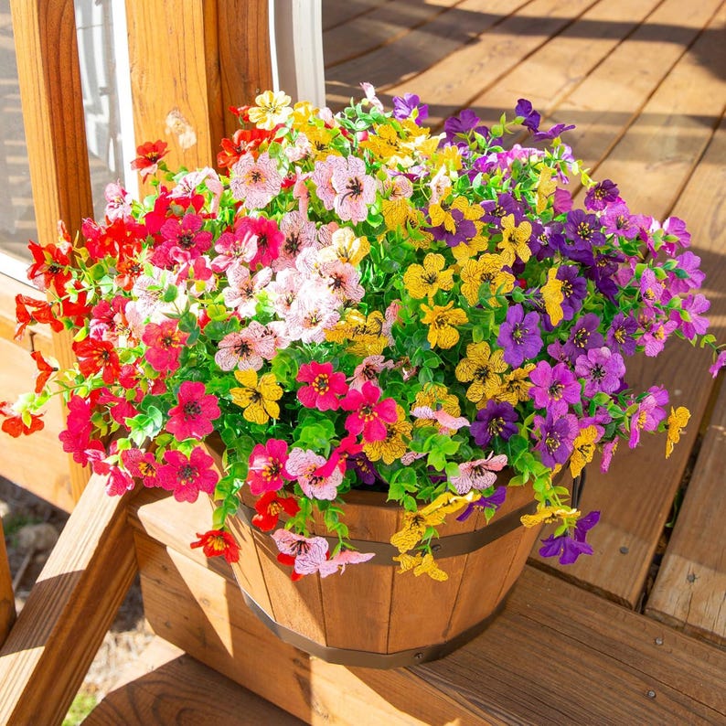 May include: A wooden barrel planter filled with a dense arrangement of artificial flowers in vibrant shades of red, pink, yellow, and purple. The flowers feature small, five-petalled blossoms with green foliage, creating a full, rounded display.