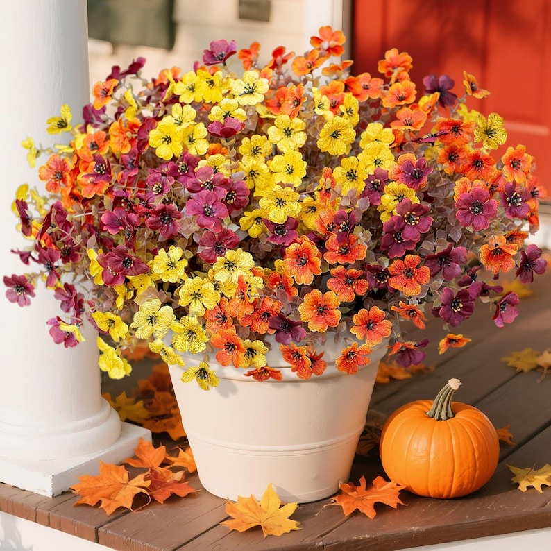 May include: A large, round, light-coloured planter filled with a dense arrangement of artificial flowers in vibrant shades of yellow, orange, and deep purple. A small, smooth, orange pumpkin sits on the surface next to the planter.
