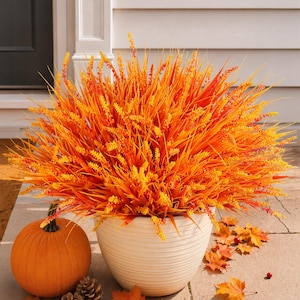 May include: A vibrant artificial plant with orange and yellow grass-like blades and small flower spikes, housed in a round, ribbed white planter. An orange pumpkin, two pinecones, and scattered autumn leaves are arranged around the base.