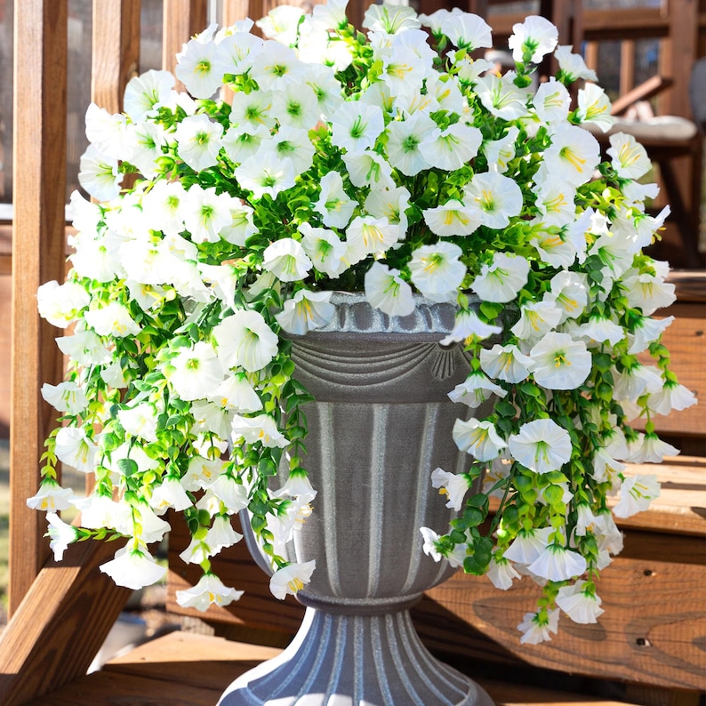 Pu&ograve; includere: Un grande vaso decorativo grigio traboccante di fiori bianchi e fogliame verde. Il vaso ha un design classico con strisce verticali e si trova su un terrazzo di legno. I fiori ricadono sui lati.