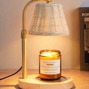 May include: A candle warmer lamp with a matte gold metal arm, a light-colored woven wicker shade, and a round natural wood base. A glass jar candle labeled Aroma Sensation Nectarine Blossom & Honey sits on the base, illuminated by the lamp's light.