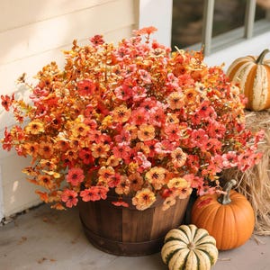 May include: A large, rounded arrangement of artificial flowers in shades of orange, yellow, and red, displayed in a dark brown wooden barrel. The floral display is surrounded by three pumpkins, including two orange ones and one striped white and green pumpkin.
