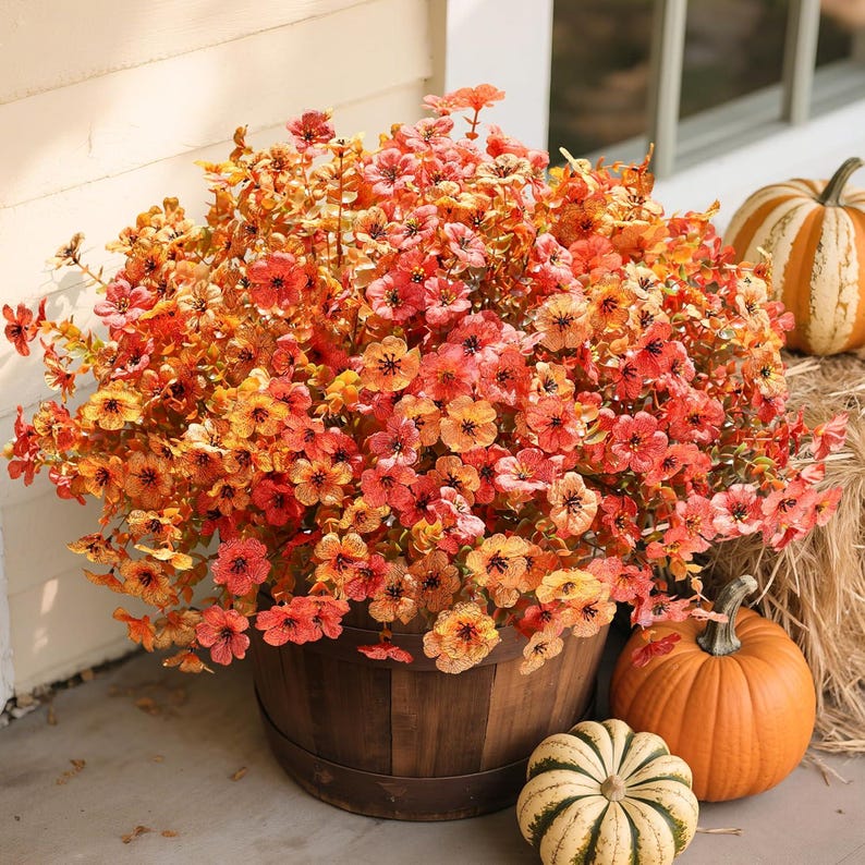 May include: A large, rounded arrangement of artificial flowers in shades of orange, yellow, and red, displayed in a dark brown wooden barrel. The floral display is surrounded by three pumpkins, including two orange ones and one striped white and green pumpkin.