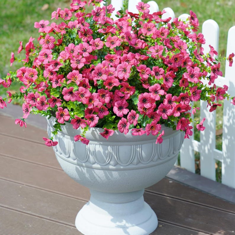 May include: A large, light grey pedestal planter filled with a dense arrangement of vibrant pink flowers. The planter features a rounded bowl shape with a decorative scalloped rim detail and a sturdy, wide base, holding numerous small blossoms.