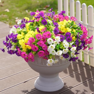 May include: A white ceramic planter filled with a vibrant mix of pink, yellow, and purple flowers. The planter has a classic, ornate design and sits on a wooden surface.