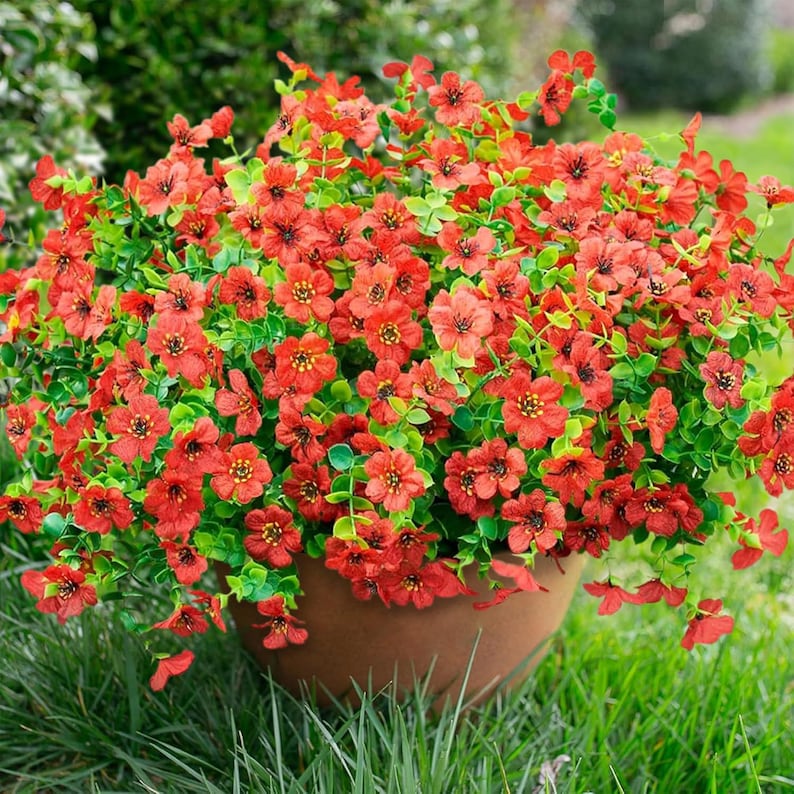 May include: A dense cluster of small, vibrant orange-red flowers with yellow centres and dark stamens, interspersed with bright green leaves. The flowers are arranged in a rounded shape and are contained within a simple, smooth, light brown pot.