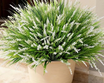 May include: A large, rounded artificial plant featuring dense, vibrant green grass blades interspersed with numerous delicate, upright white flower spikes, all contained within a smooth, light beige, circular ceramic planter pot.