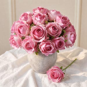 May include: A bouquet of pink roses in a textured gray vase. The roses have multiple layers of petals and are in full bloom. One rose lies separately on a white cloth. The arrangement is a classic floral display.