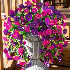Pu&ograve; includere: Un'urna grigia e ornata traboccante di vivaci fiori artificiali. I fiori sono un mix di viola intenso e rosa brillante, con centri gialli e fogliame verde. La composizione &egrave; ambientata su uno sfondo di legno, creando un'esposizione colorata.
