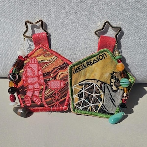 May include: Two pentagonal fabric keychains with star-shaped metal clips. The left one features a red astronaut embroidery on a swirling orange and brown background. The right one shows a person near a geometric dome with the text LIFE IS REASON.