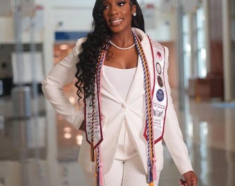 May include: A woman in a white pantsuit and a white top, wearing a pearl necklace and a graduation sash. The sash has multiple medallions and tassels. The background is a hallway with architectural details.