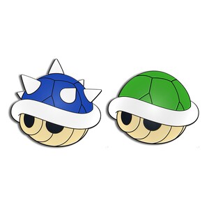 May include: Two cartoon turtle shells, one blue with white spikes and the other green. Both shells have a white rim and a beige underside with black eye-like shapes, set against a white background.