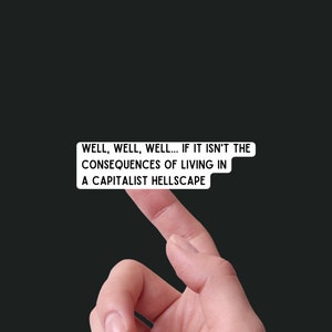 May include: A white sticker with black text that reads: "WELL, WELL, WELL... IF IT ISN'T THE CONSEQUENCES OF LIVING IN A CAPITALIST HELLSCAPE." The sticker is being held by a finger against a black background.
