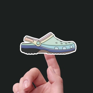 May include: A colorful, multi-colored, cartoon-style illustration of a clog shoe. The shoe is a light blue, green, and pink color with a black sole. The shoe is being held up by a finger.