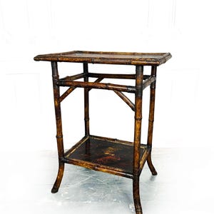 May include: A vintage bamboo side table with two tiers. The table has a dark brown finish with a rectangular top and a lower shelf. The legs are curved, and the table has a classic, antique aesthetic. The table measures approximately 61 cm tall.