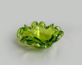 May include: A green glass ashtray with a wavy, organic design. The ashtray is made of clear glass with a green tint.