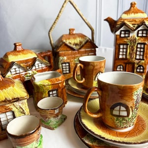 Puede incluir: Un juego de té vintage con temática de cabaña con tetera, jarra de crema, azucarero, tazas, platillos y hueveras. El juego está hecho de cerámica y presenta colores marrón, amarillo y verde. Las piezas están decoradas con imágenes de cabañas y techos de paja.