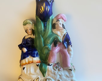 May include: A vintage ceramic figurine of a couple sitting beside a blue and green tulip. The woman is wearing a blue dress with a white apron and a green hat. The man is wearing a blue jacket, pink pants, and a white hat. The figurine is decorated with gold accents.