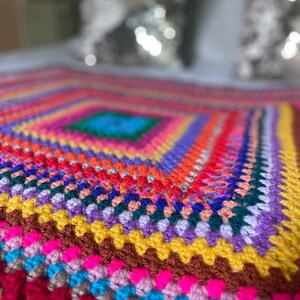 Puede incluir: Una manta de crochet colorida con un patrón cuadrado en el centro. La manta está hecha con una variedad de colores brillantes, incluyendo rojo, naranja, amarillo, verde, azul, morado y rosa.