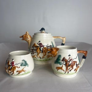 May include: A vintage ceramic tea set with a fox head teapot and two mugs. The teapot and mugs are decorated with a hunting scene featuring riders on horseback and hounds chasing a fox.