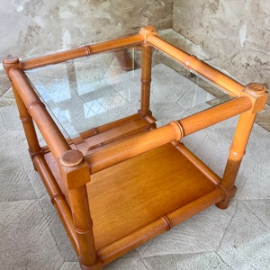 May include: A square, two-tiered end table with a glass top and a lower wooden shelf. The table is made of light brown wood with a bamboo-like design.