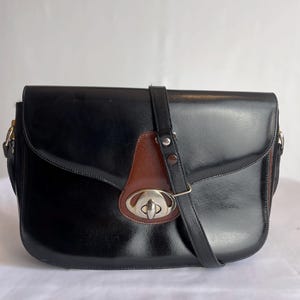 May include: A black leather satchel with a brown leather flap and a silver clasp. The bag has a long adjustable strap.