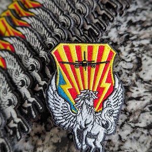 May include: A stack of embroidered patches featuring a white winged horse with a red, yellow and blue shield behind it. The shield has a stylised airplane inside.