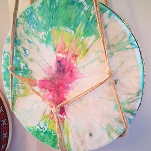 May include: A round, tie-dye drum with a vibrant pattern of pink, green, and blue hues. The drum is suspended by a natural fiber rope, creating a decorative and functional piece. The design is reminiscent of a watercolor painting.