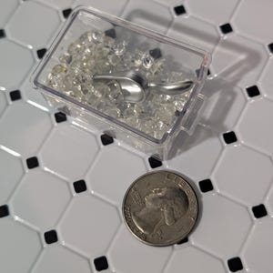 Puede incluir: Un recipiente de plástico transparente lleno de cuentas claras y facetadas y una pequeña cuchara plateada. Una moneda está al lado del recipiente. El recipiente está sobre una superficie de baldosas blancas con detalles cuadrados negros.