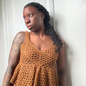 May include: A sleeveless, mustard-brown crochet top featuring a scoop neckline and a flared peplum hem. The garment is constructed with an open-work, grid-like stitch pattern throughout the bodice and a slightly denser, vertical stitch design at the waist.