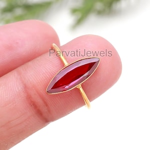 May include: A gold ring featuring a marquise-cut, deep red gemstone. The ring's slender band and setting are gold-colored, creating a striking contrast with the vibrant red stone. The ring is held up by a finger.