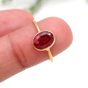 Pode incluir: Um anel de ouro com uma vibrante pedra de rubi de corte oval. O rubi está engastado numa delicada banda de ouro, criando um design clássico e elegante. O anel é exibido sobre um fundo suave e desfocado.