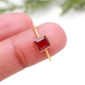 Op de afbeelding: Een goudkleurige ring met een vierkante, dieprode edelsteen. De ring heeft een dunne band en de edelsteen is gezet in een eenvoudig ontwerp. De ring wordt in een hand gehouden, met de tekst "Parva Jewels" zichtbaar.