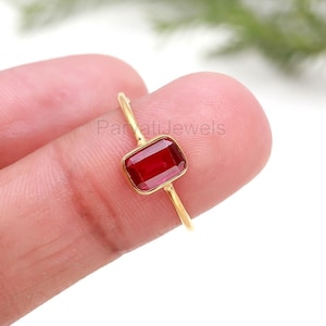 Pode incluir: Um anel de ouro com uma pedra preciosa retangular, vermelho escuro. O anel tem uma banda fina e delicada e uma moldura simples. A pedra preciosa tem aproximadamente 0,8 cm de comprimento e 0,5 cm de largura. O anel é exibido contra um fundo desfocado.