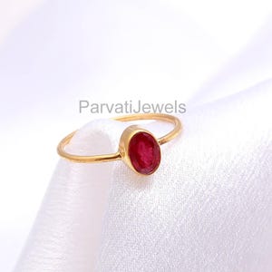 May include: A gold ring with a ruby gemstone. The ring features a thin gold band and an oval-shaped, deep red ruby. The ring is set against a white, textured background. The text "ParvatiJewels" is visible in the background.