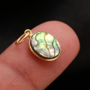 May include: A gold-colored pendant with an oval abalone shell center. The shell displays iridescent green, blue, and purple hues. The pendant has a small loop for a chain. The text "harvati jewels" is visible on the pendant.