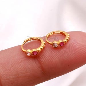 May include: Two small, gold-colored hoop earrings. Each earring features a clear and a red gemstone. The earrings are displayed on a finger against a white background. The earrings have a textured design around the hoop.