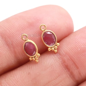 May include: A pair of gold-plated earrings, each with an oval ruby gemstone. The earrings have a small loop at the top and a cluster of small beads at the bottom. The earrings are presented against a light background, highlighting their design.
