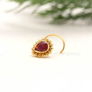 May include: A gold nose ring featuring a pear-shaped red gemstone at the center, surrounded by a decorative beaded border. The jewelry piece has a curved wire stem and includes the text PARVATI JEWELS printed faintly near the base.