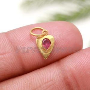 May include: A gold heart-shaped pendant with a pink gemstone in the center. The pendant has a loop at the top for attaching to a chain.