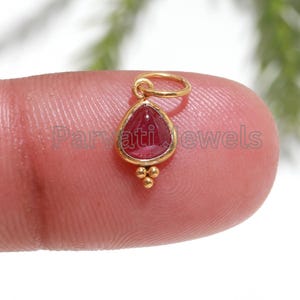 Op de afbeelding: Een gouden neusring met een rode edelsteen. De ring heeft de vorm van een traan en heeft een kleine, decoratieve bal aan de onderkant.