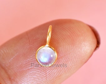 May include: A gold pendant with a round, iridescent moonstone charm. The pendant is on a finger.