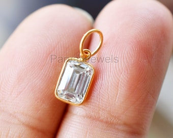 Puede incluir: Un colgante de oro con una piedra preciosa transparente de talla esmeralda. El colgante está en un dedo.