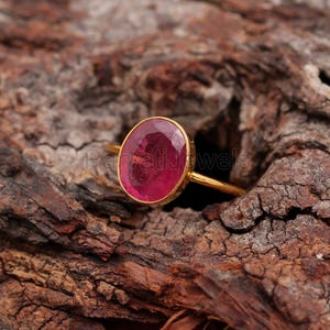 May include: A gold ring featuring a large, oval-shaped, pink gemstone. The ring is displayed on a wooden surface.