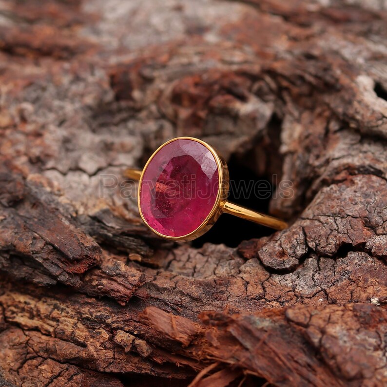 May include: A gold ring featuring a large, oval-shaped, pink gemstone. The ring is displayed on a wooden surface.