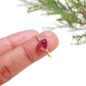 May include: A gold ring featuring a triangular, deep pink gemstone. The ring is held by a person. The text "Parvati Jewels" is visible on the image.