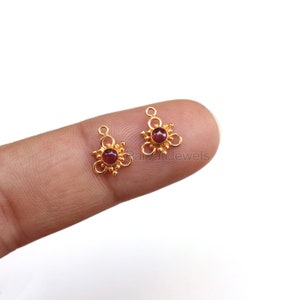 Pode incluir: Dois pingentes dourados com uma gema vermelha no centro de cada um. Cada pingente tem um laço na parte superior para prender a um colar ou pulseira.